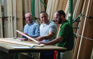 3 men standing at a large table in a woodworking shop looking at a long wooden profile piece